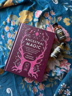 A book titled 'Ancestral Magic' by Frankie Castanea lies on a blue floral blanket, styled with two small amber bottles. The book cover is deep purple with pink tree illustrations and white text.