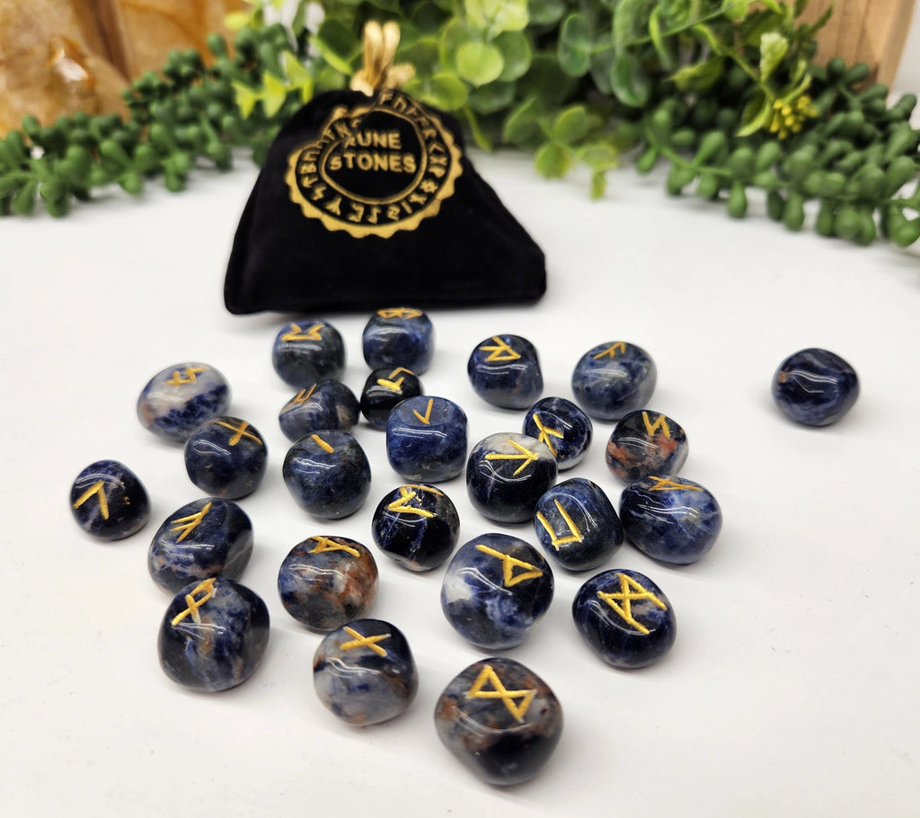 Set of blue stones with gold symbols next to a black bag labeled 'Nune & Stones' on a white surface with greenery in the background.
