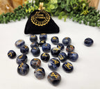 Set of blue stones with gold symbols next to a black bag labeled 'Nune & Stones' on a white surface with greenery in the background.