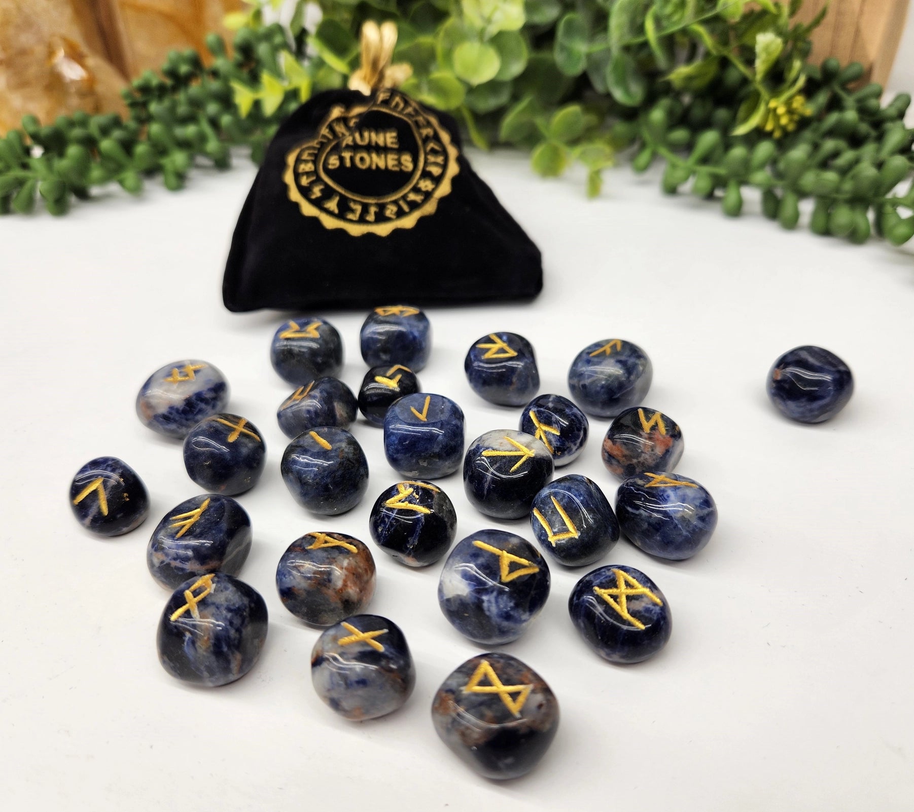 Set of blue stones with gold symbols next to a black bag labeled 'Nune & Stones' on a white surface with greenery in the background.