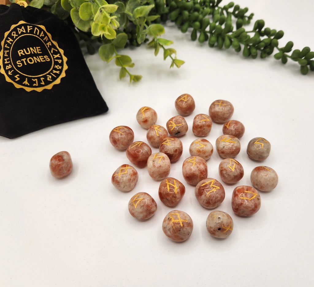 Rune stones with gold runes on a white surface, next to a black bag labeled 'Rune Stones' and greenery.